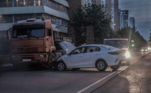 A car is damaged after a collision with a semi-trailer truck. After a semi-truck crash, first steps include getting medical care and protecting evidence from the scene.