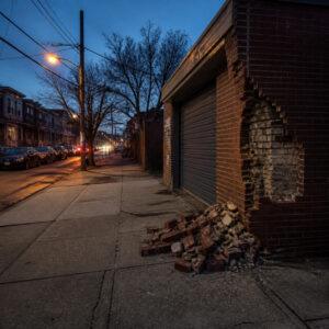 Fallen bricks on a city sidewalk beside an old brick garage wall